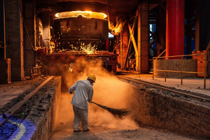 A worker in a yellow helmet sweeps dust near molten metal and sparks inside a steel factory.