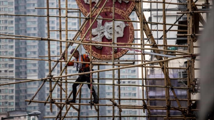 A scaffolder climbs bamboo scaffolding being constructed around a large neon sign, with high-rise buildings in the background.