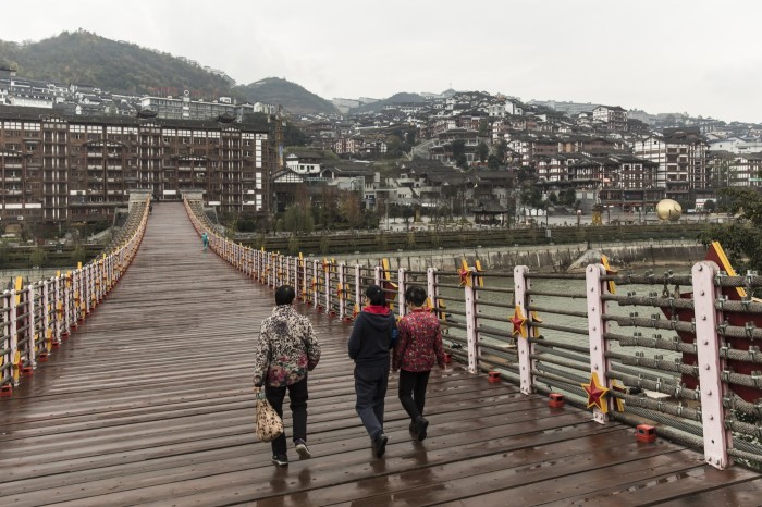 Three women walk on a wooden bridge decorated with red and yellow stars, with Maotai town’s hillside buildings visible ahead