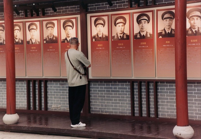 A man stands in front of a display of red-framed text and portraits of men in military uniform.
