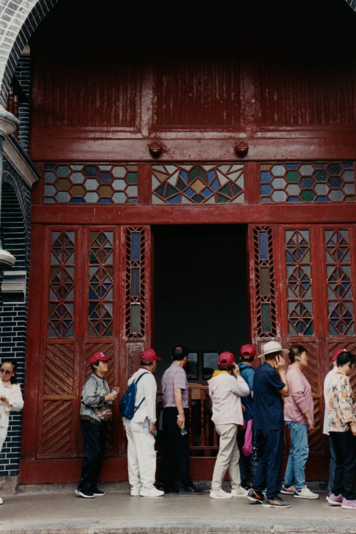A line of people, variously dressed, at the ornate red-painted doorway of a building.