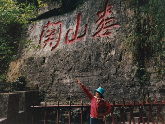 A person in a red top and blue hat poses in front of a cliff face etched with giant Chinese letters in red.