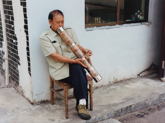 A man seated outdoors on a small wooden chair next to a whitewashed wall smoking through a large tube.