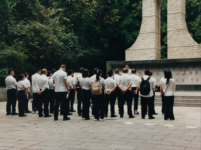 A group of mostly men in white shirts and black trousers stand in front of a modern-looking memorial structure.