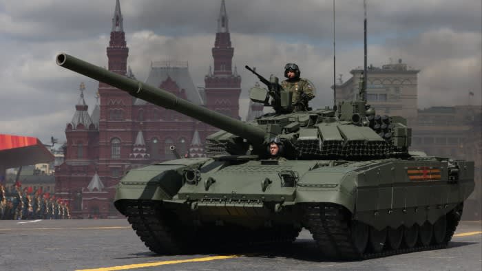 A Russian tank with two soldiers visible passes through Moscow’s Red Square during a Victory Day military parade, with historic red buildings in the background.