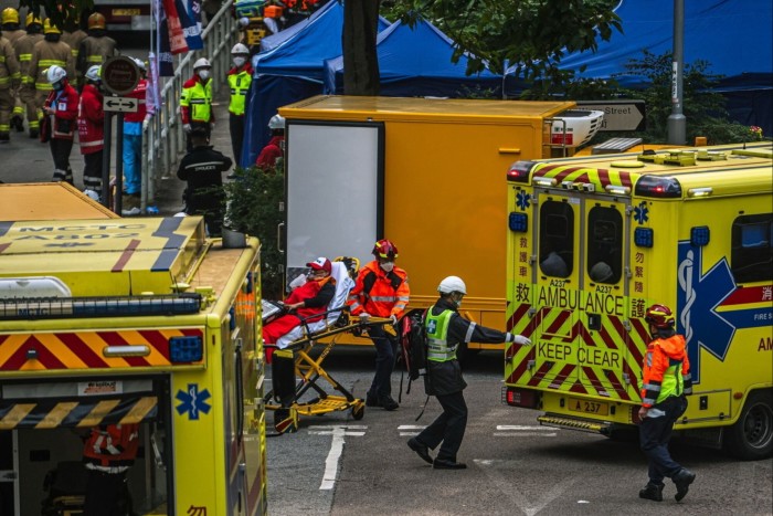 Rescue workers move a person on a stretcher towards an ambulance near several emergency vehicles and tents