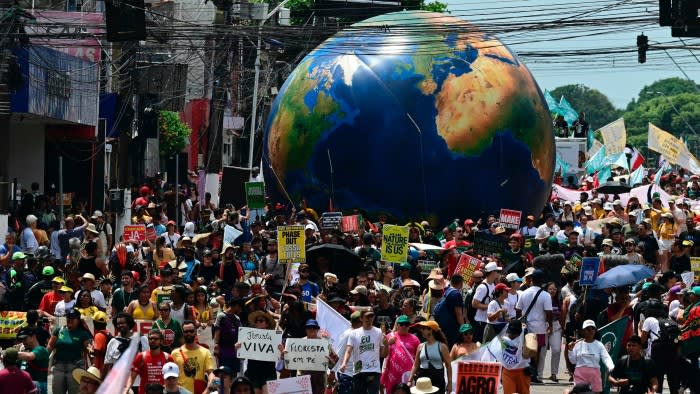 A large crowd marches down a street carrying protest signs and banners, with a giant inflatable Earth globe in the background.