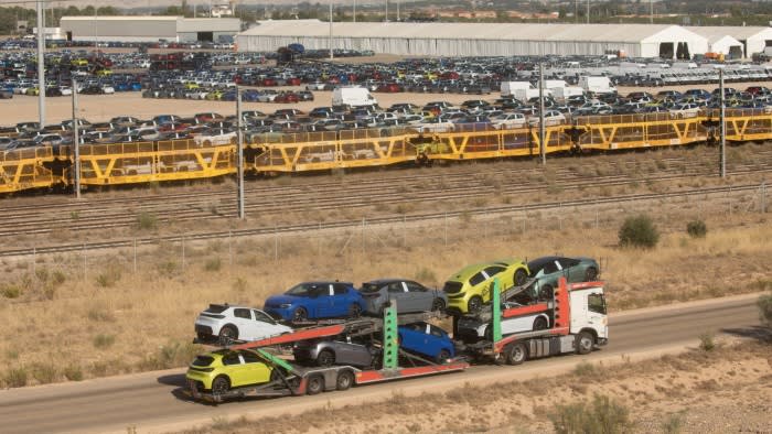 Rows of new cars fill a large parking lot at a Stellantis site in Zaragoza, Spain, with a car carrier truck and yellow rail cars used for vehicle transport in the foreground.