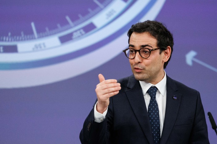 Stéphane Séjourné gestures while speaking at a press conference, standing in front of a large graphic display.