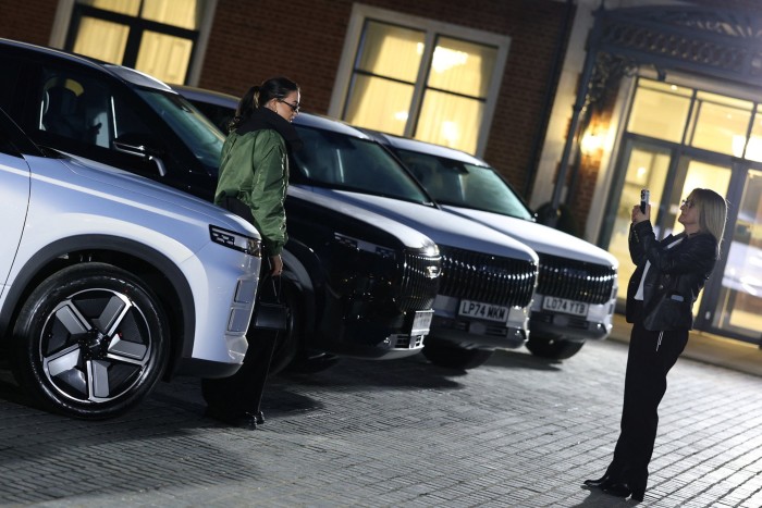 A guest poses beside a white Jaecoo 7 SUV while another person takes her photo at a launch event with several Jaecoo 7 vehicles lined up.