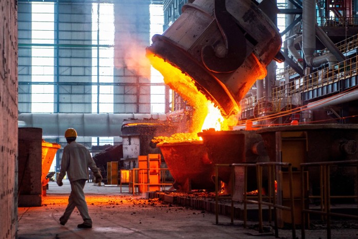 A worker in a hard hat walks past molten steel pouring from a large container inside a steel factory.