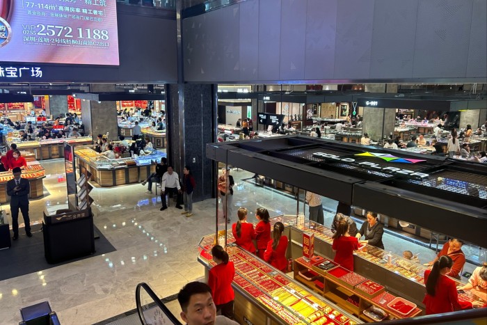 A busy gold jewellery market in Shuibei, with multiple glass display counters and staff in red uniforms assisting customers.