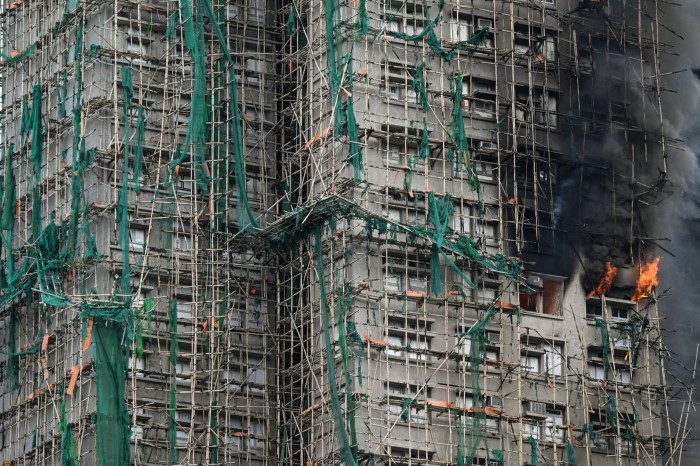 Flames and thick smoke pour from windows of a high-rise apartment block covered in bamboo scaffolding and green netting.