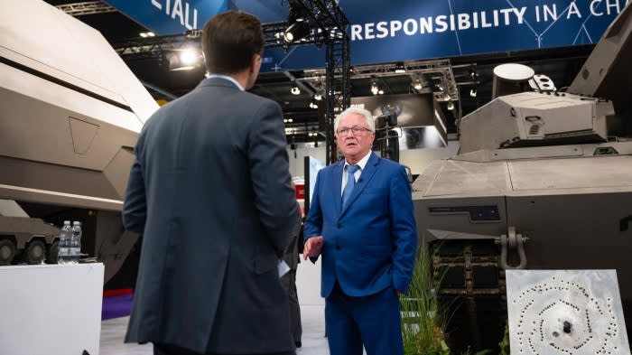 Armin Papperger speaks during a television interview at the DSEI 2025 exhibition, standing near armoured vehicles