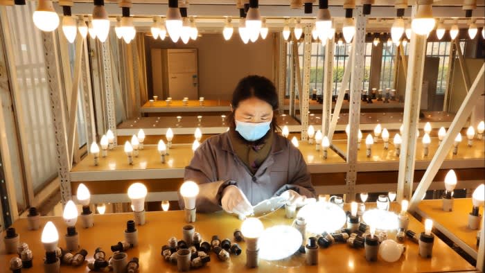 An employee wearing a face mask assembles LED energy-saving lamps on a brightly lit factory production line.
