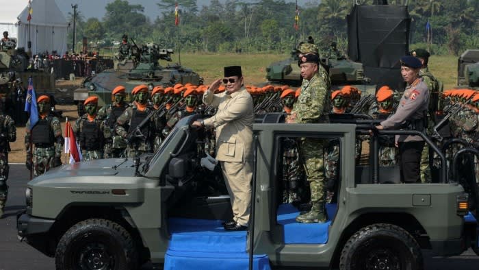 President Prabowo Subianto salutes troops while standing in a vehicle during a military ceremony, with soldiers and armoured vehicles in the background.