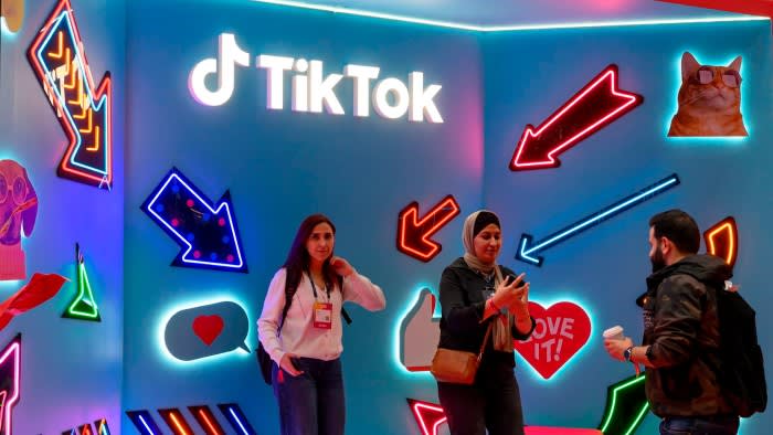 Three visitors stand and talk at the TikTok booth, decorated with neon arrows and internet-themed icons, at the Web Summit.