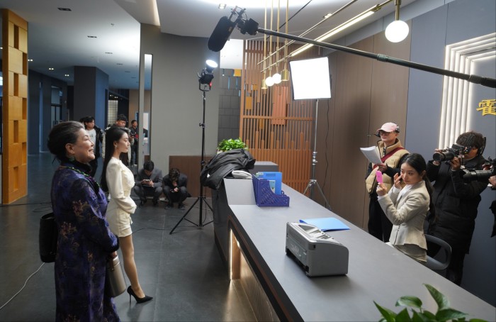 Cast and crew film a scene at a modern office set, with lighting and sound equipment visible and several people preparing for the shoot.