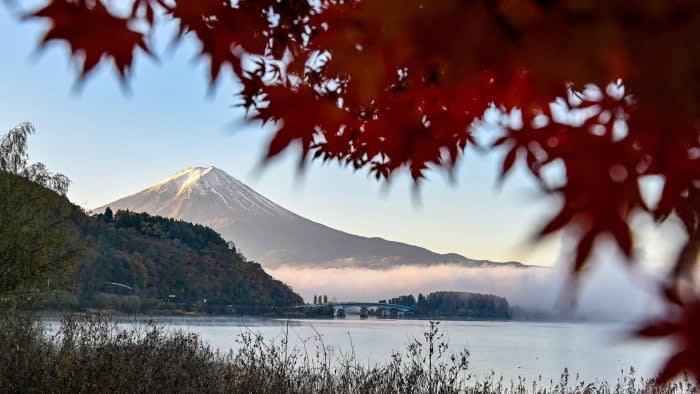 Mount Fuji with a snow-capped peak is seen in the distance, framed by vivid red autumn leaves and a calm lake in the foreground.