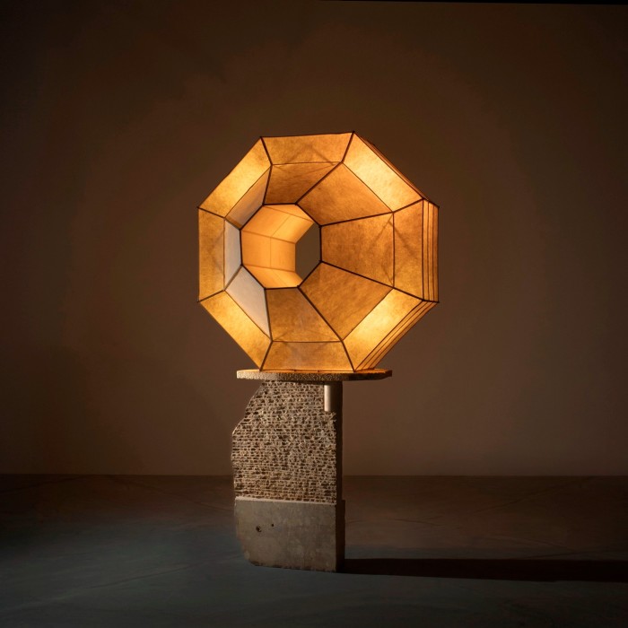 A geometric floor lamp with a glowing, faceted shade sits atop a textured stone and concrete base.