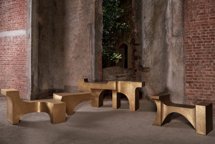 Three sculptural benches and one table from Vikram Goyal's Golden Arc Collection, featuring gold-toned, geometric forms with arched bases, set in a weathered brick and concrete space.