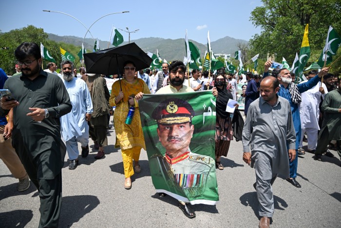 A man holds a large portrait of Munir during a rally, with others waving Pakistani flags in the background.