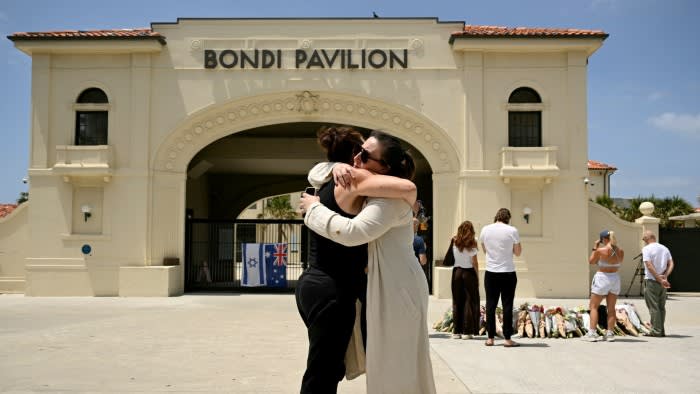Two mourners embrace in front of Bondi Pavilion while others stand near floral tributes and Israeli and Australian flags.
