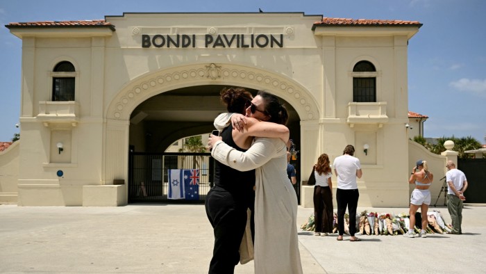Two mourners embrace in front of Bondi Pavilion while others stand near floral tributes and Israeli and Australian flags.