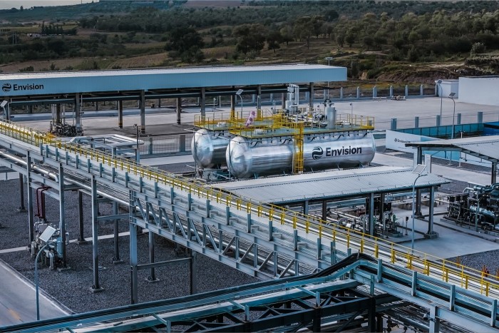 Industrial facility with large cylindrical tanks labeled "Envision," surrounded by metal walkways, pipes, and a partially covered structure.
