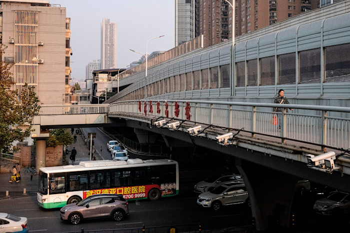 A city bus travels on a street below an elevated pedestrian walkway in Wuhan, with several surveillance cameras mounted along the bridge.