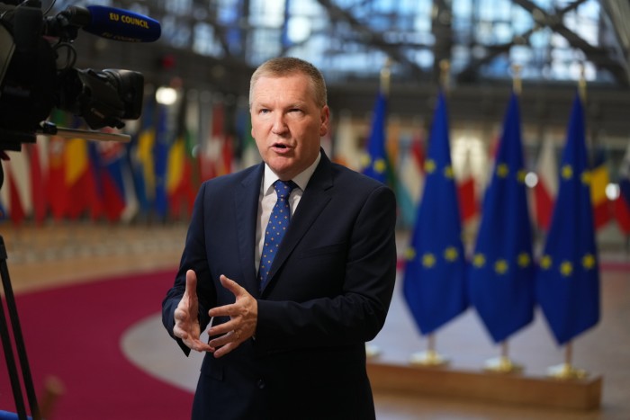Michael McGrath speaks to media in front of EU flags and national flags at the European Council building.