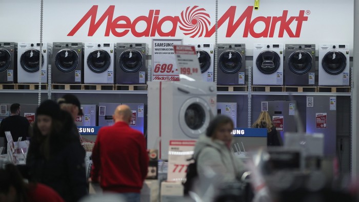 Customers look at washing machines on display beneath a large MediaMarkt sign inside an electronics store.