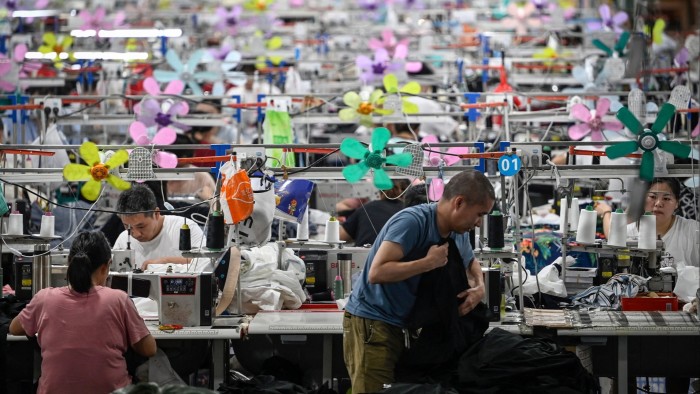 Rows of workers operate sewing machines at a busy textile factory, with colourful fans attached above each workstation