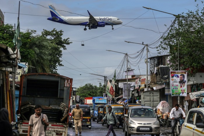 An IndiGo airplane descends above a busy street with cars, rickshaws, pedestrians and market stalls in Mumbai