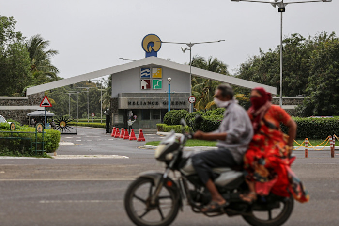 Entry gate to Reliance Greens township with “Reliance Greens” signage, as two people on a motorcycle pass by in the foreground.