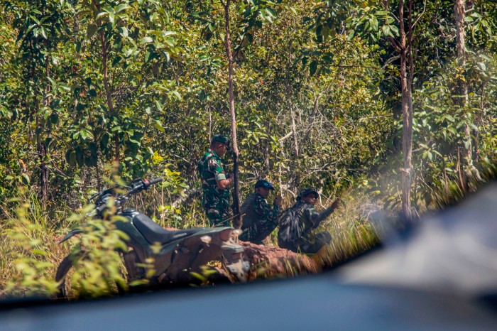 Three soldiers are next to a motorcycle against a backdrop of dense foliage