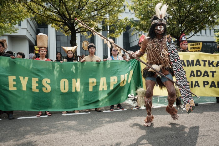A man is visible in indigenous dress and holding a spear while banners and protesters can be seen in the background
