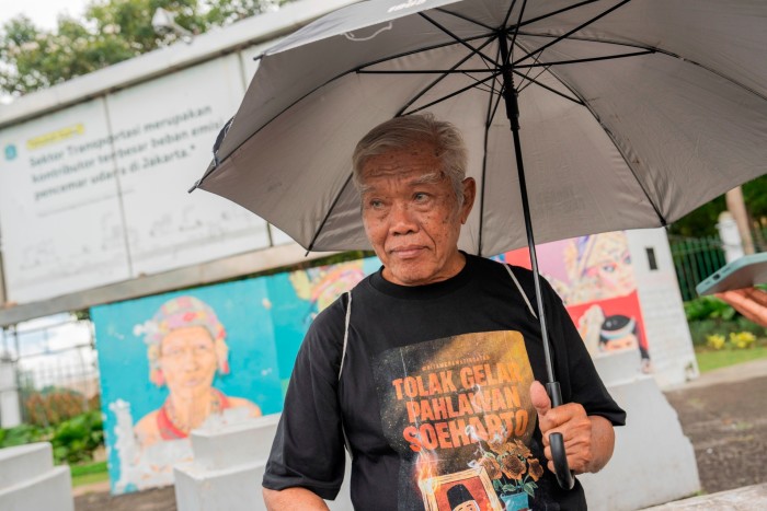 Bedjo Untung stands under an umbrella, wearing a black T-shirt with anti-Suharto text, in front of murals in Jakarta.