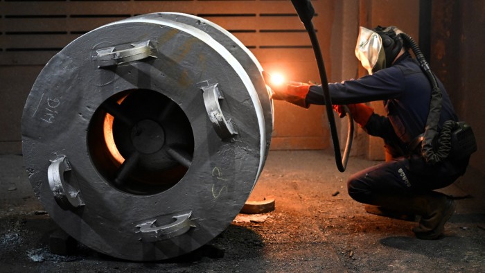 A worker in protective gear welds a large metal component, with sparks illuminating the foundry floor.