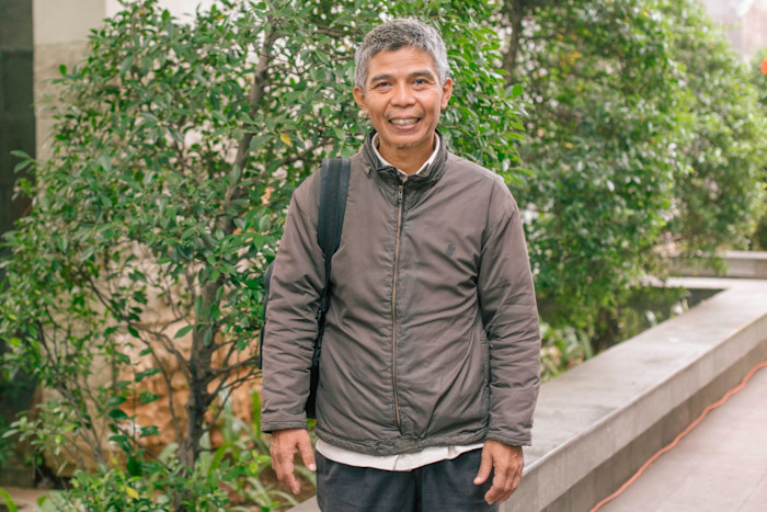 Hidayat stands outdoors with a backpack, smiling, wearing a brown jacket in front of greenery.