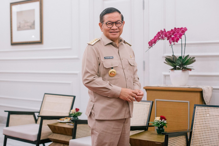 Pramono Anung stands smiling in his official uniform in an office, with chairs and a potted orchid visible behind him.
