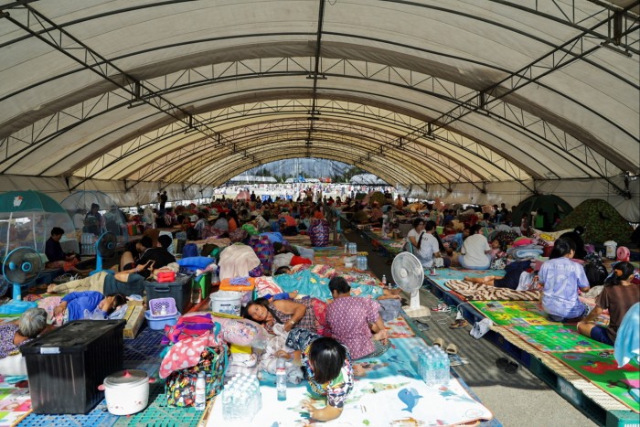 People rest and lie on mats under a large temporary shelter in Thailand, surrounded by personal belongings, after fleeing military clashes