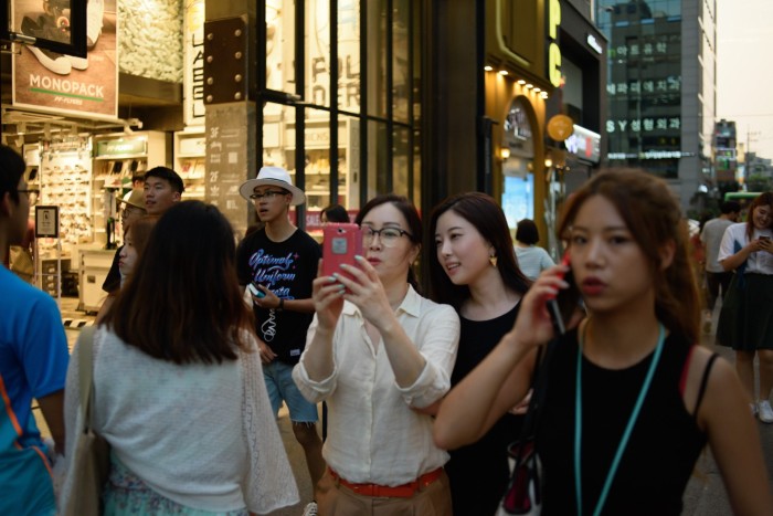 Two women stand close together on a busy Seoul street, looking at a mobile phone, surrounded by pedestrians.