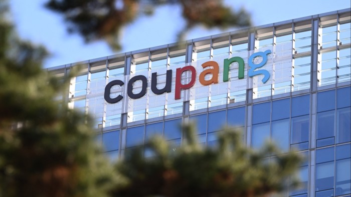 The Coupang logo is displayed on the upper facade of the company’s glass headquarters building in Seoul, partially obscured by tree branches.