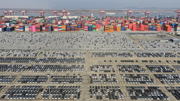 Rows of new vehicles lined up in a large lot at Nanjing Port, with colorful shipping containers and cranes in the background.