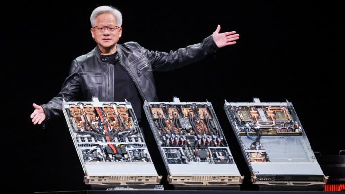 Jensen Huang gestures with open arms while standing behind three open computer server units displaying internal hardware