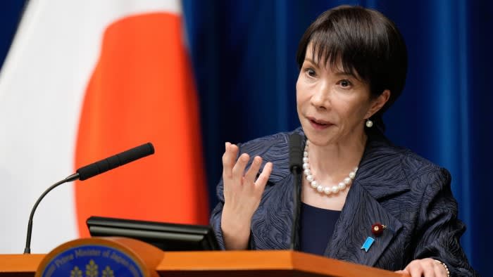 Sanae Takaichi speaks at a podium during a press conference, gesturing with her hand. A Japanese flag is visible in the background.