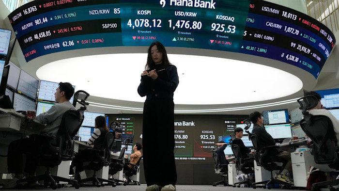 A currency trader stands with a notepad as others work at computer terminals beneath electronic boards displaying financial data at Hana Bank.