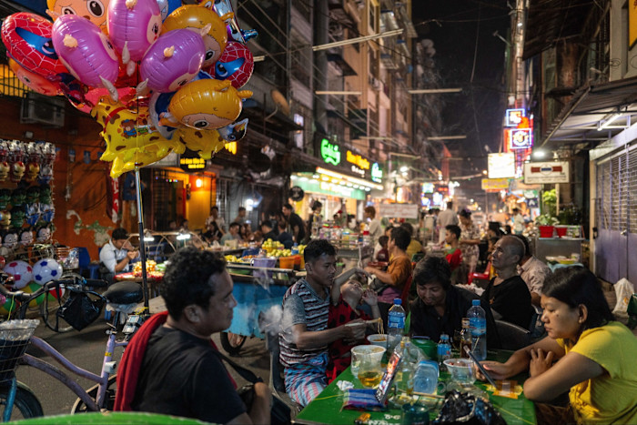 People sit at green tables eating and drinking at a busy open-air Chinatown eatery at night, with colorful balloons and neon signs nearby.