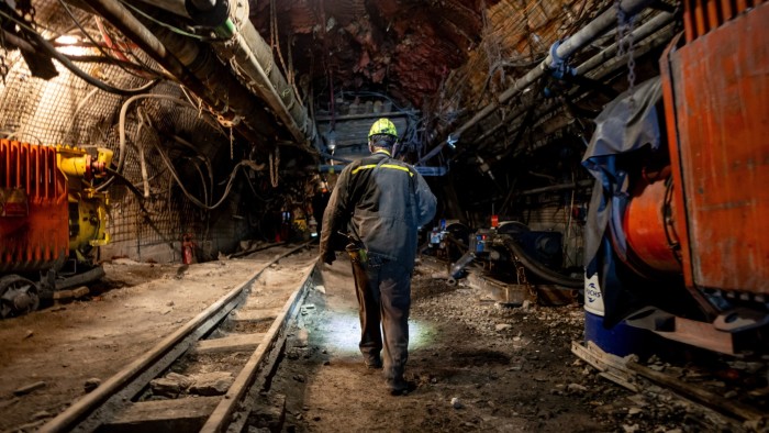 A coal miner wearing a helmet and work clothes walks along a tunnel deep underground at the CSM coal mine in Stonava, surrounded by mining equipment and machinery.